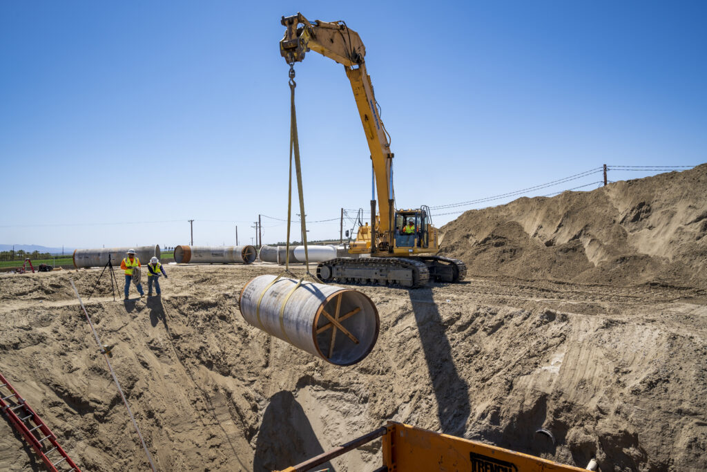 Workers guide the placement of a large pipeline section at the Oasis In-Lieu Recharge Phase 2 Project in Thermal, California.